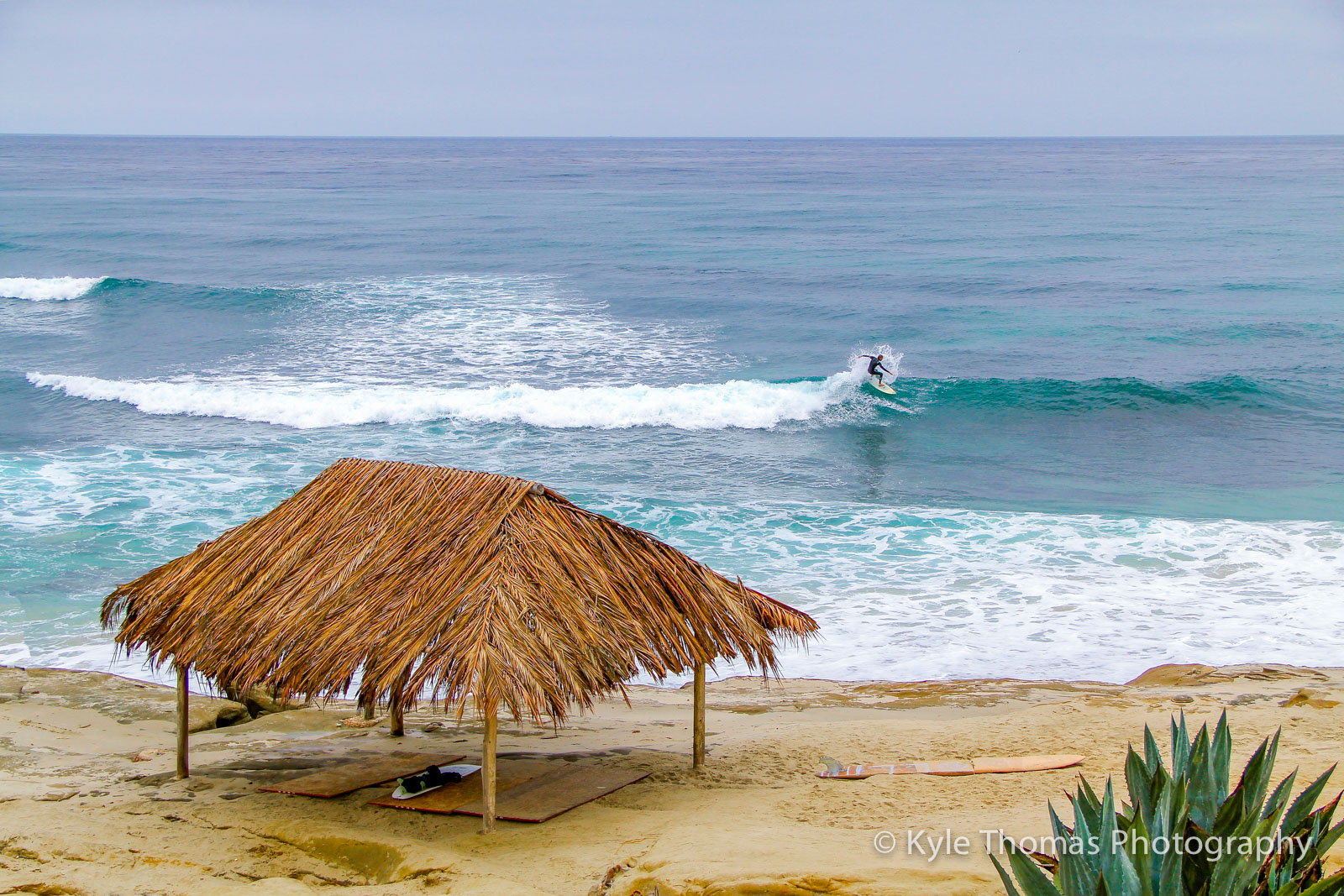 Thatched palapa on the sand above the surf line, surfer in the water beyond