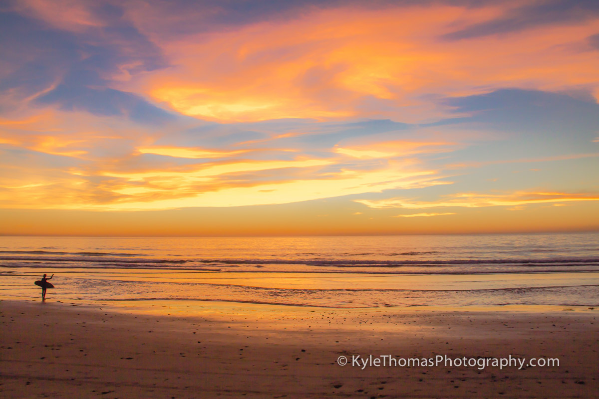 Surfers silhouetted against an orange and violet sunset over the Pacific