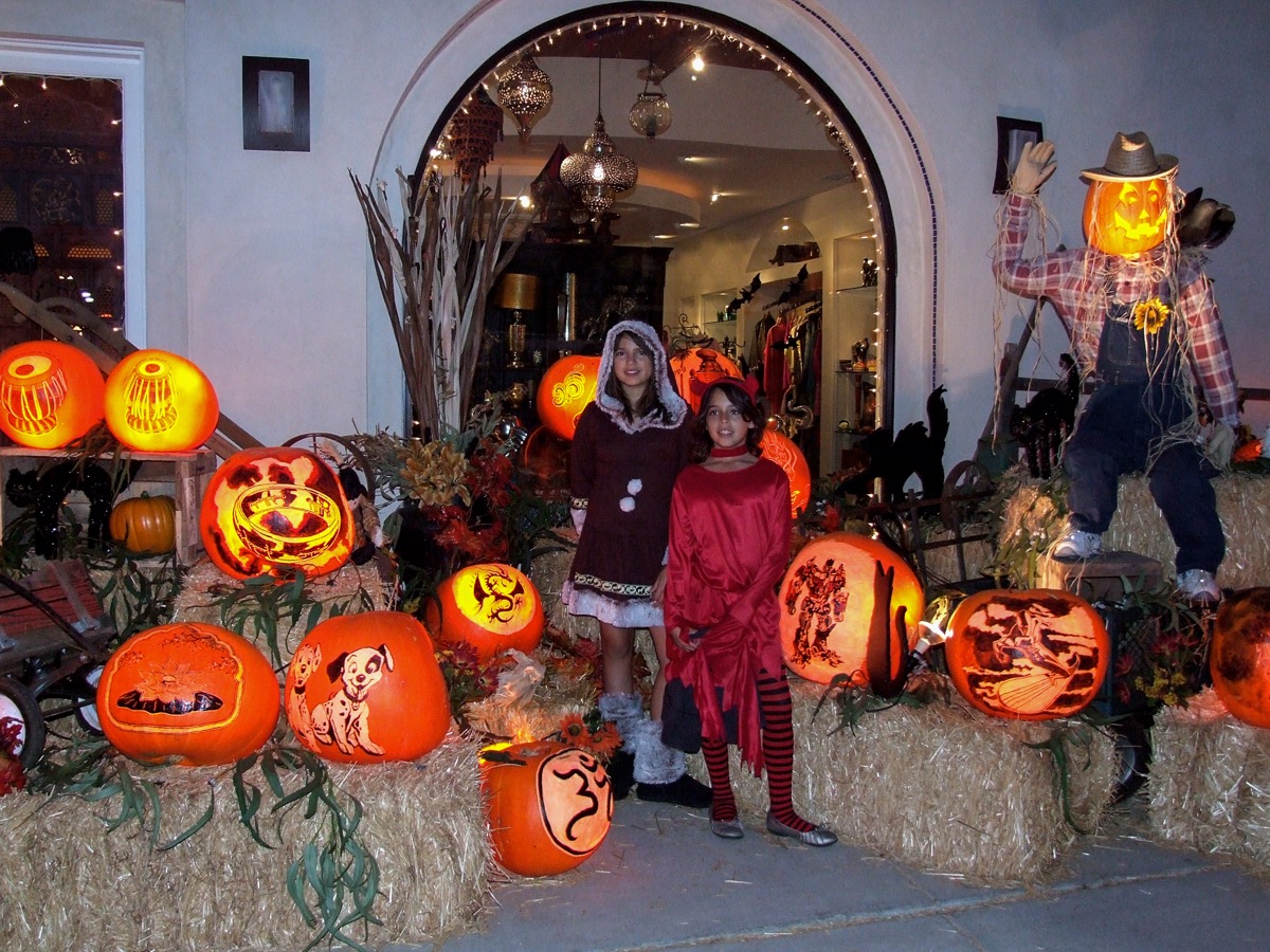 A shop doorway on Coast Highway 101 banked with straw bales and dozens of lit carved pumpkins, a scarecrow with a jack-o-lantern head above, two children in costume standing in the middle of the display