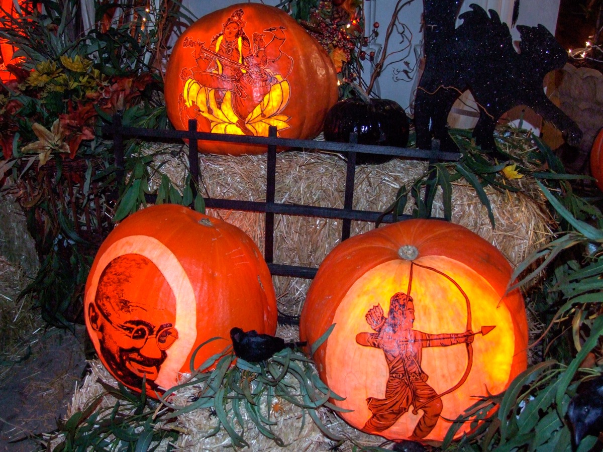 Three lit pumpkins on straw bales, one carved with the face of Mahatma Gandhi, one with an archer drawing a bow, one above with a figure seated on a lotus
