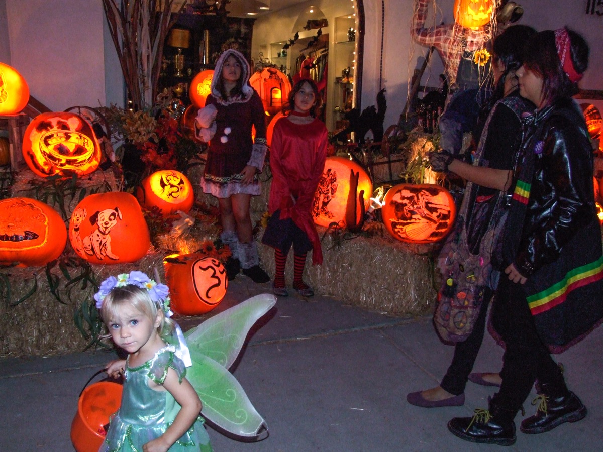 Trick-or-treaters on the sidewalk in front of a lit pumpkin display, a small girl in a fairy costume with a pumpkin pail in the foreground, older children in costume behind her