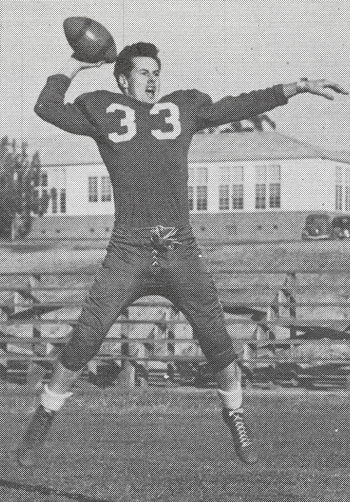 A young football player in a dark jersey numbered 33 leaping with the ball cocked back to pass, school buildings behind him