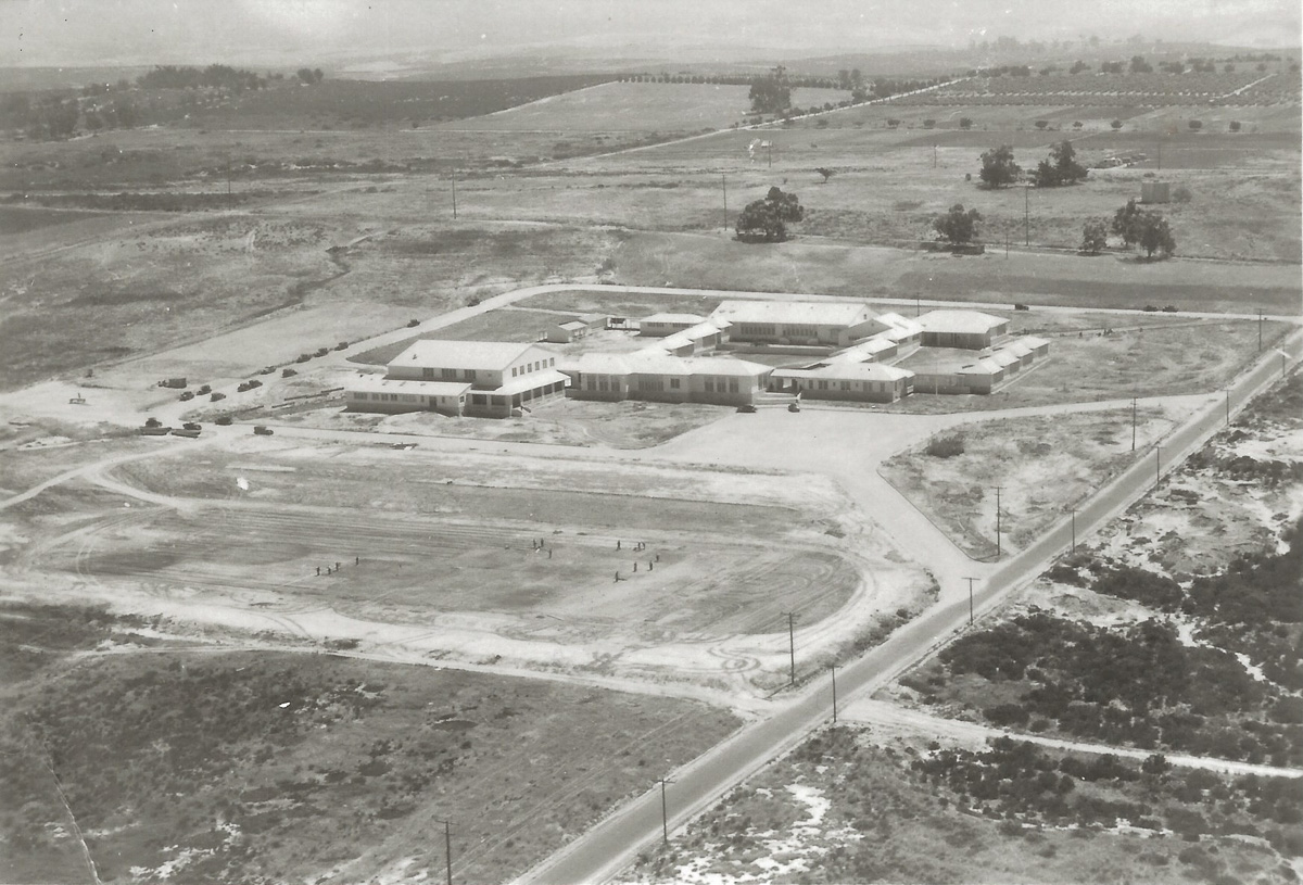 An aerial photograph of the new school buildings standing alone on open, scrubby land, a dirt athletic field in front with a few figures on it, and a single paved road cutting diagonally past