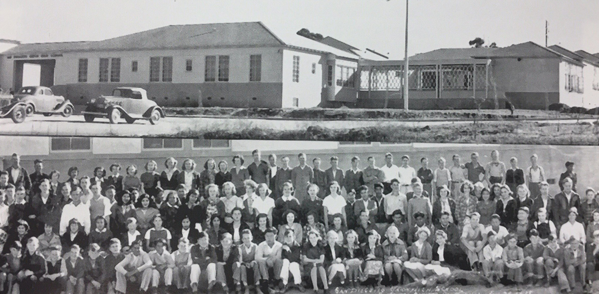 A large class of students seated and standing in rows on the grass in front of low stucco school buildings with tile roofs, two 1930s cars parked on the road behind them