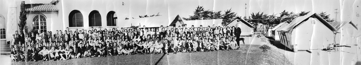 A wide panorama of the first senior class posed on a lawn beside an arched stucco building, a man in a dark suit and a dog standing at the end of the group, and a row of canvas-roofed tent classrooms under cypress trees at right