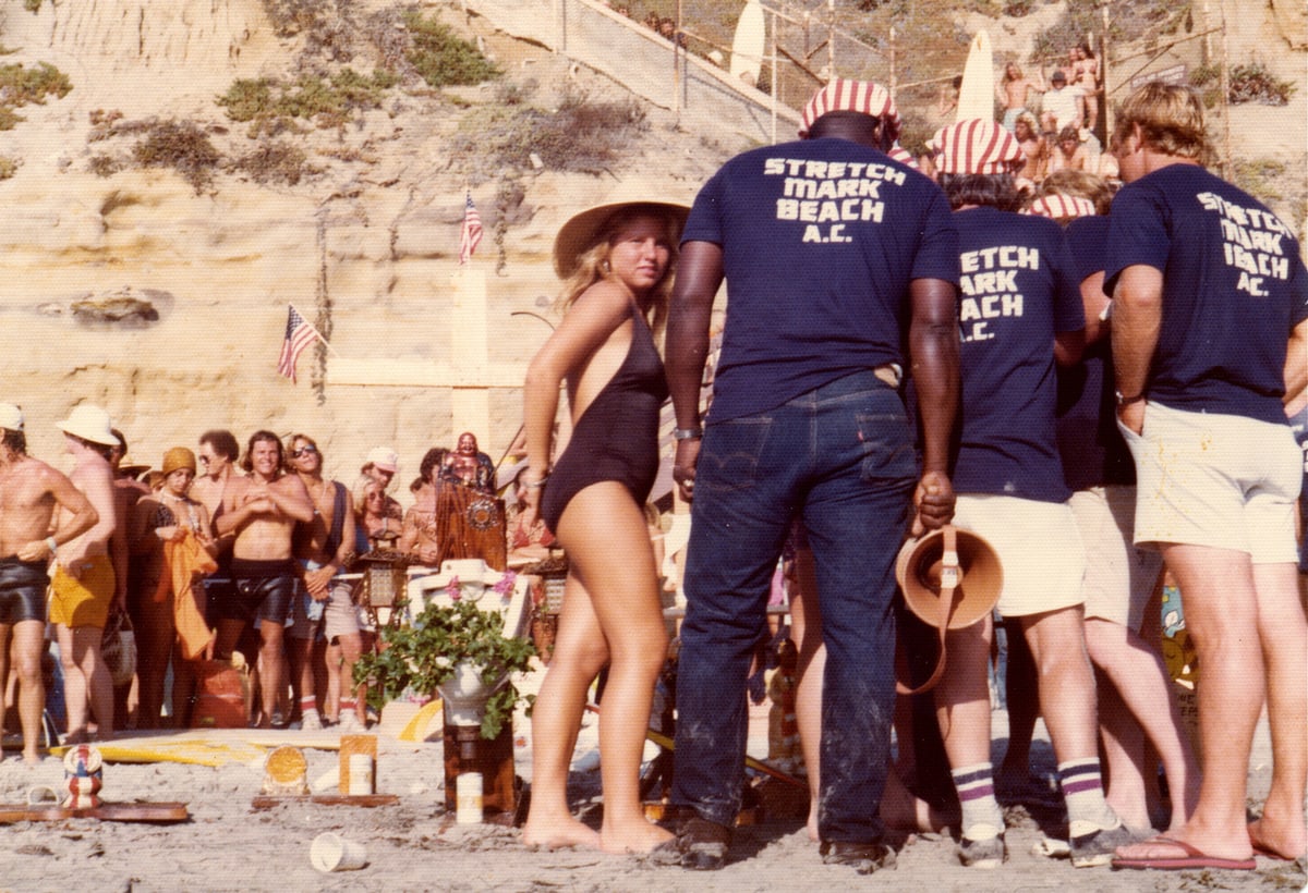 The 1975 Stonesteps Surfing Contest on the sand, a crowd around the judging table, men in Stretch Mark Beach A.C. shirts