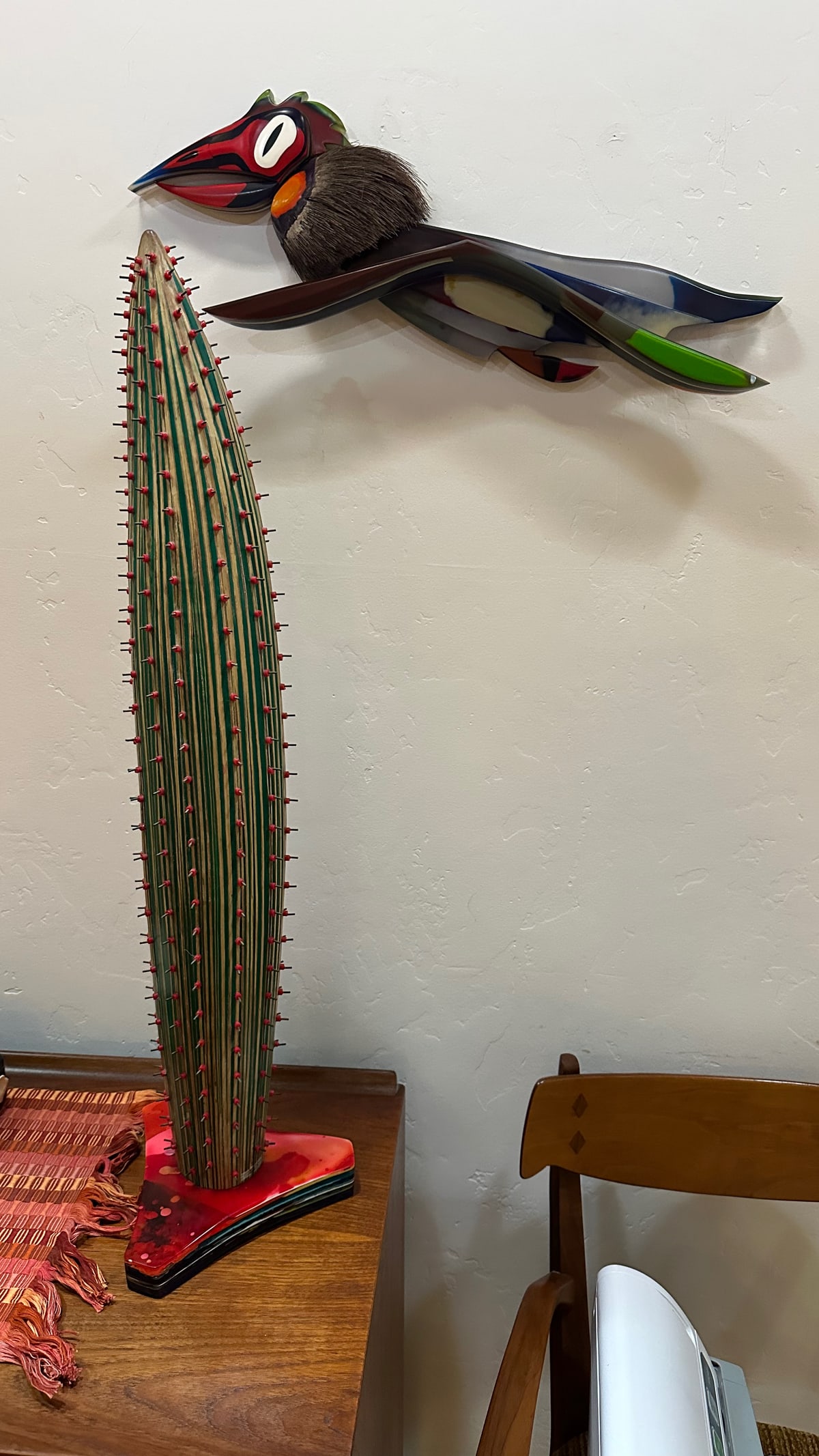 A resin bird sculpture mounted on a wall above a tall green and red cactus sculpture standing on a cabinet