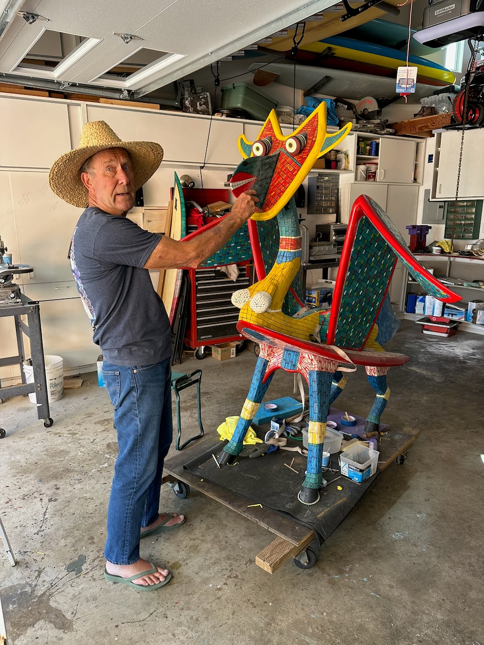 John Peugh in a straw hat working on a large mosaic-covered winged creature sculpture in his garage