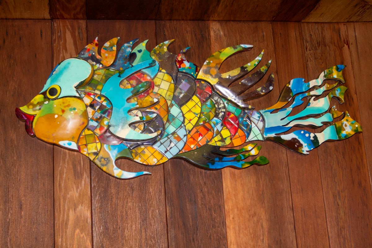 A large fish sculpture cut and assembled from multicoloured recycled surfboard resin, mounted on a wood-panelled wall