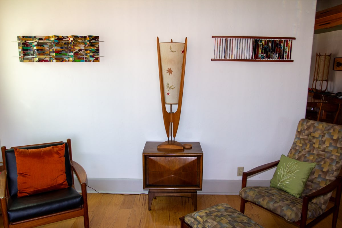 A tall wood lamp made by John Peugh standing on a credenza, with his mosaic tile sculpture hanging on the wall at left