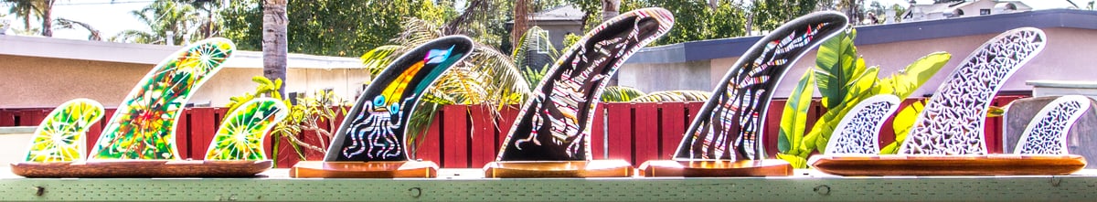 A row of seven surfboard fins made from patterned recycled resin, lined up along a rail outdoors