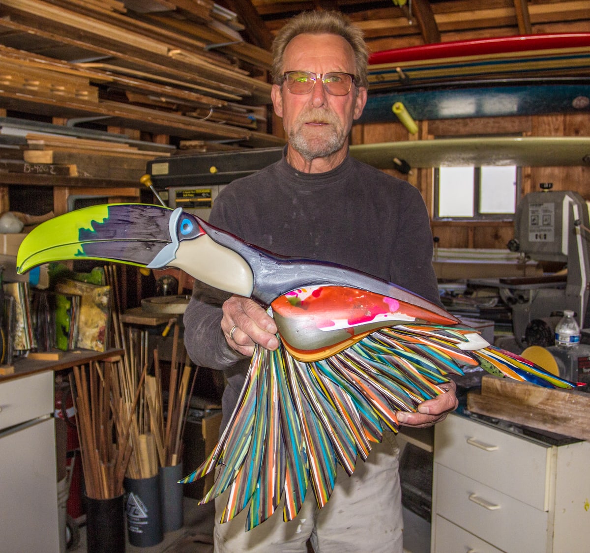 John Peugh holding a large toucan sculpture built from recycled surfboard resin in his workshop