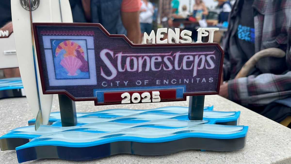 Close-up of the 2025 Stonesteps Men's first-place trophy, a layered blue resin wave base beneath a plaque and a small surfboard