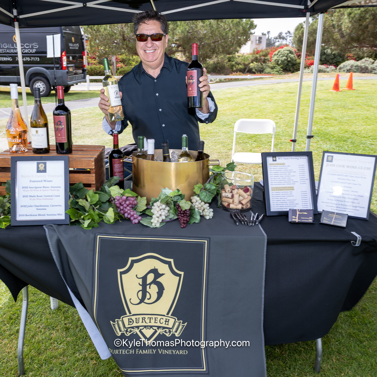James Valles in sunglasses smiling behind a vendor table under a black canopy, holding up two bottles of wine, the Burtech shield logo on the tablecloth and bottles chilling in a brass tub