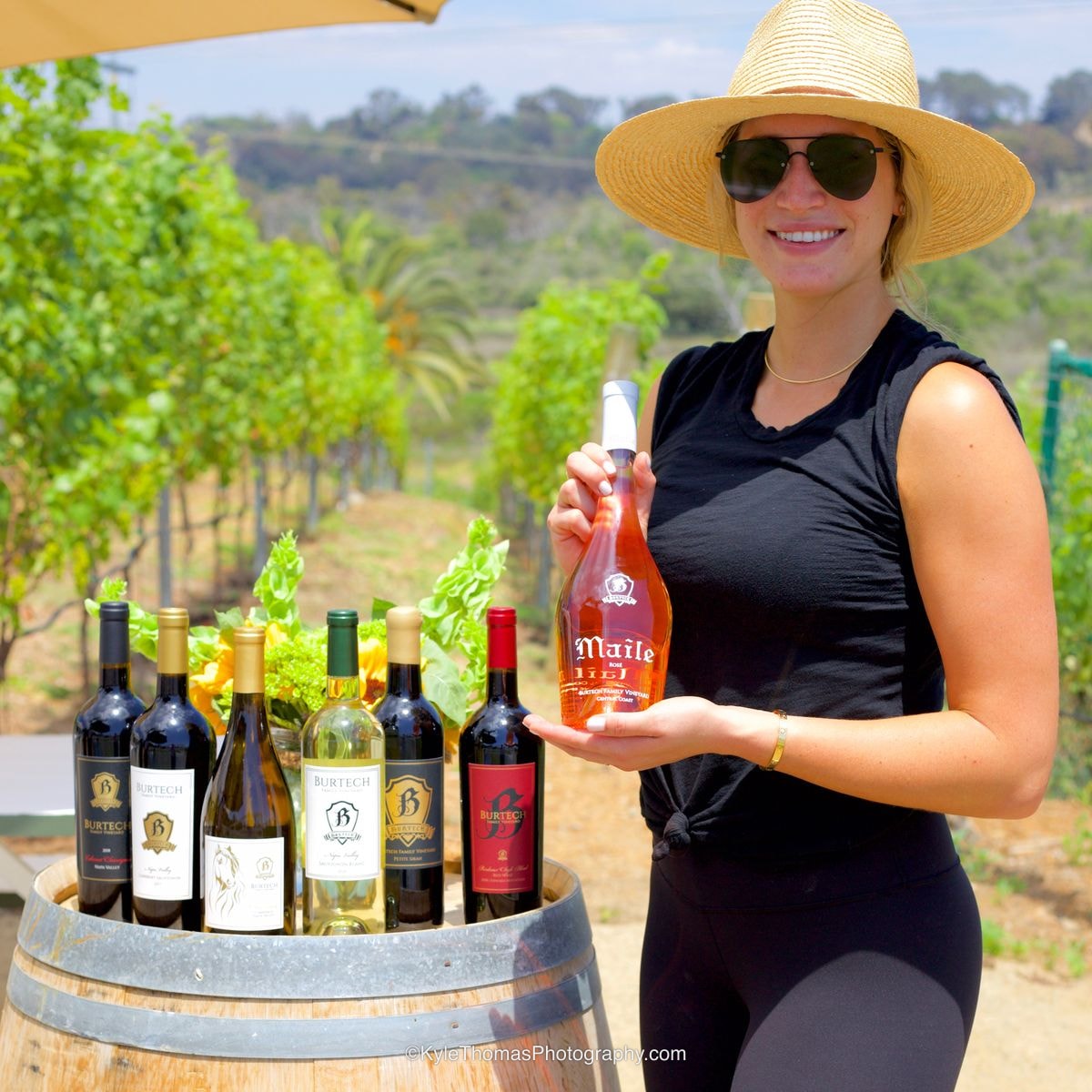 A young woman in a straw hat and sunglasses standing beside a wine barrel topped with six bottles of Burtech wine, holding up a bottle of Maile Rosé, vineyard rows and hills behind her