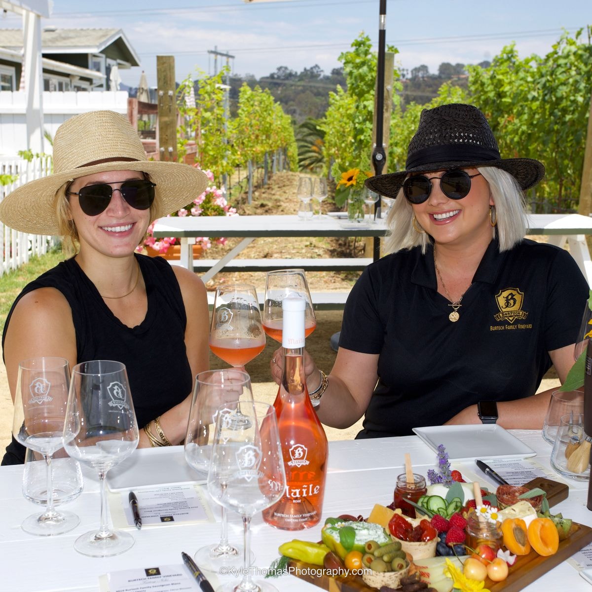 Two smiling women in sunglasses and hats seated at a white tasting table, pink rosé in their glasses, a bottle of Maile Rosé and a charcuterie board of fruit, cheese and olives in front of them