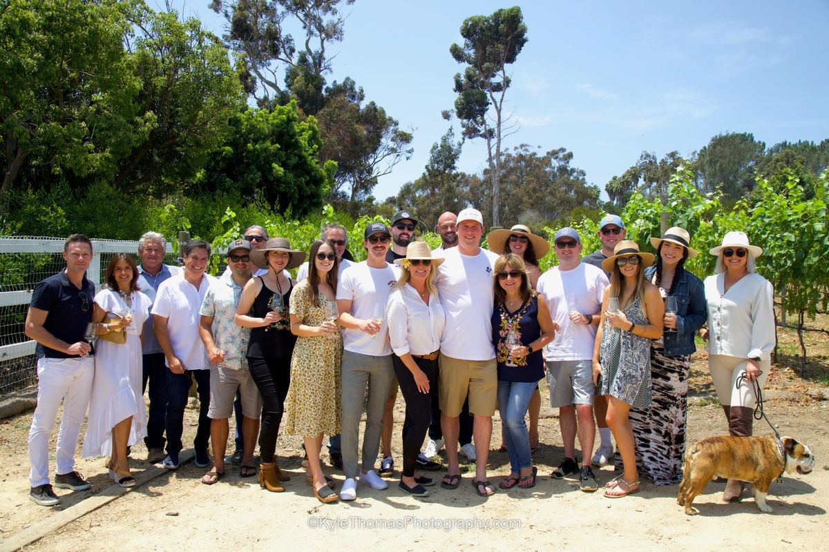 About twenty guests in sun hats and summer clothes posed together on a dirt path in front of the vines and tall eucalyptus trees, a bulldog on a leash at the end of the row
