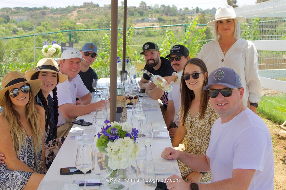 A long white table under an umbrella crowded with smiling guests in hats and sunglasses, glasses of wine and a vase of white and purple flowers down the middle
