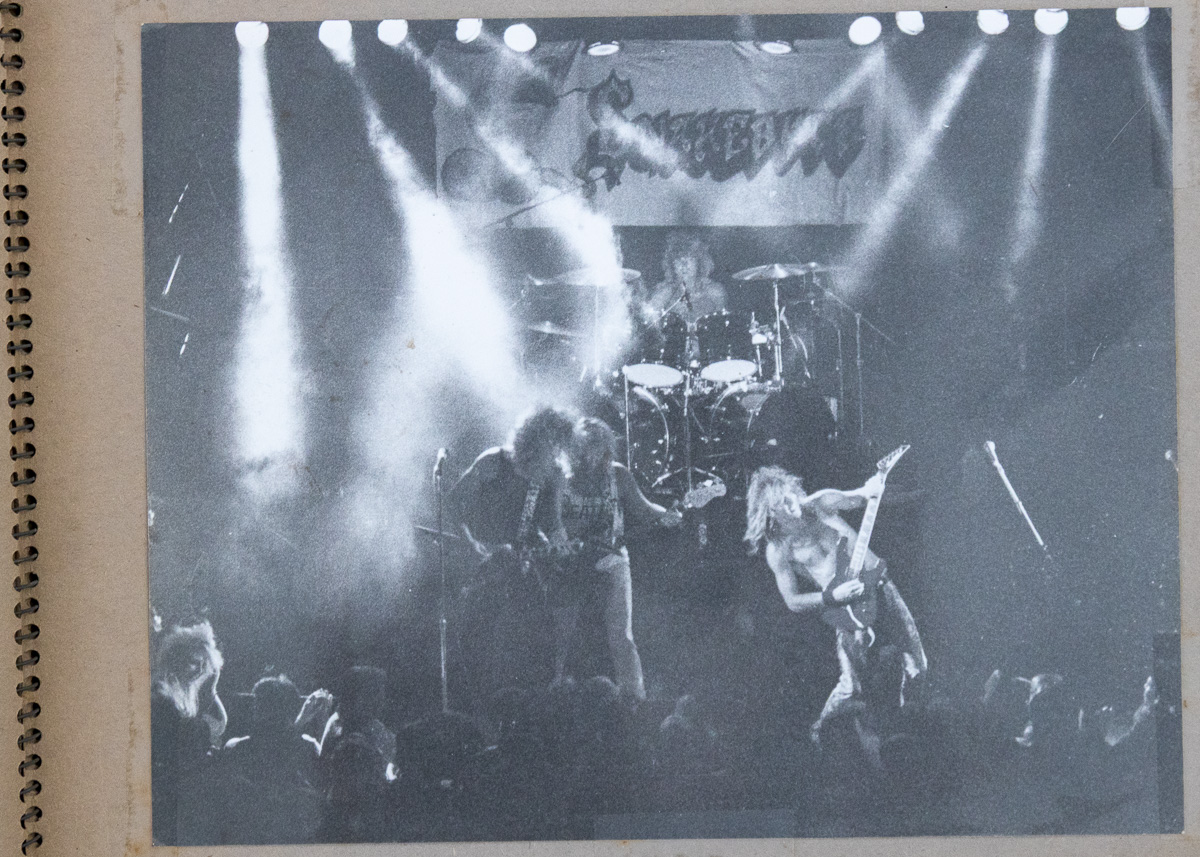 A black and white print in a scrapbook showing Snakebyte performing under stage lights beneath their banner, with the crowd in the foreground