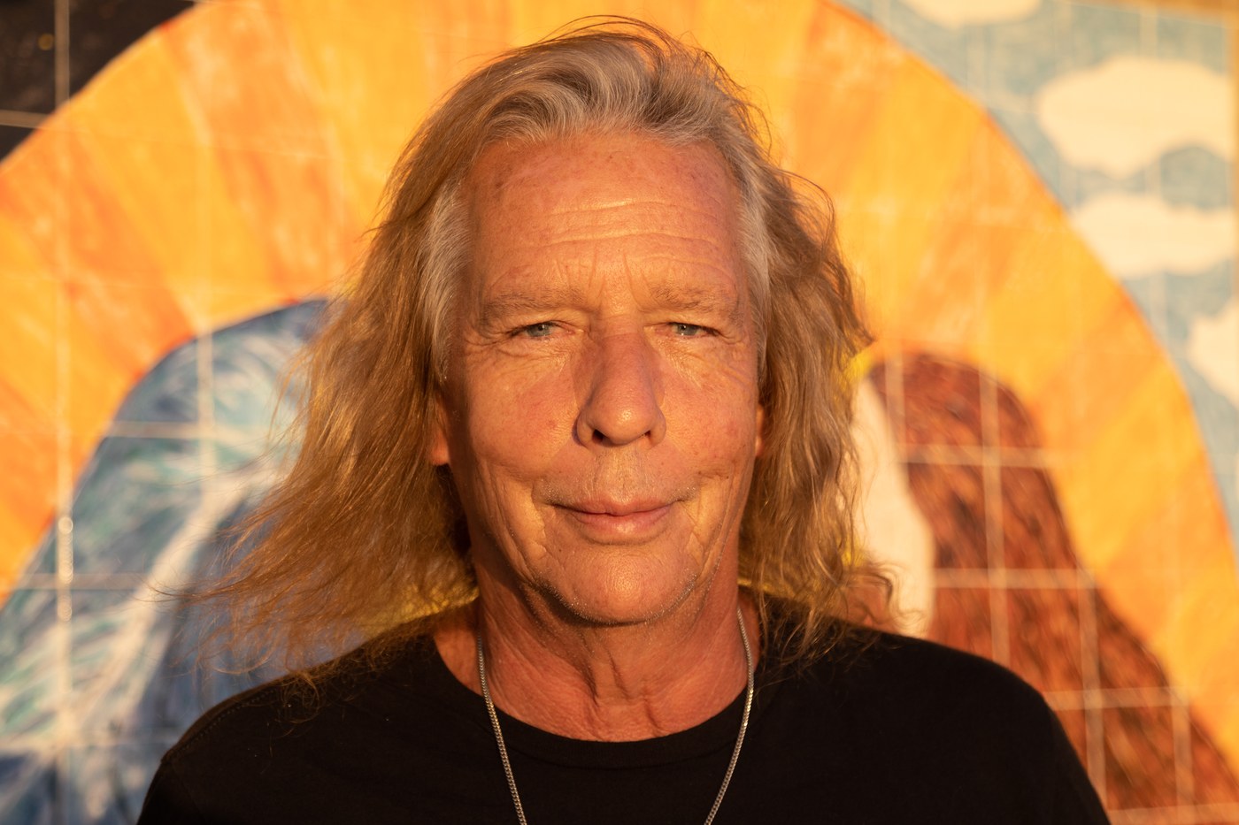 Wally Passet in 2021, long blond hair, black shirt, against an orange and blue painted backdrop