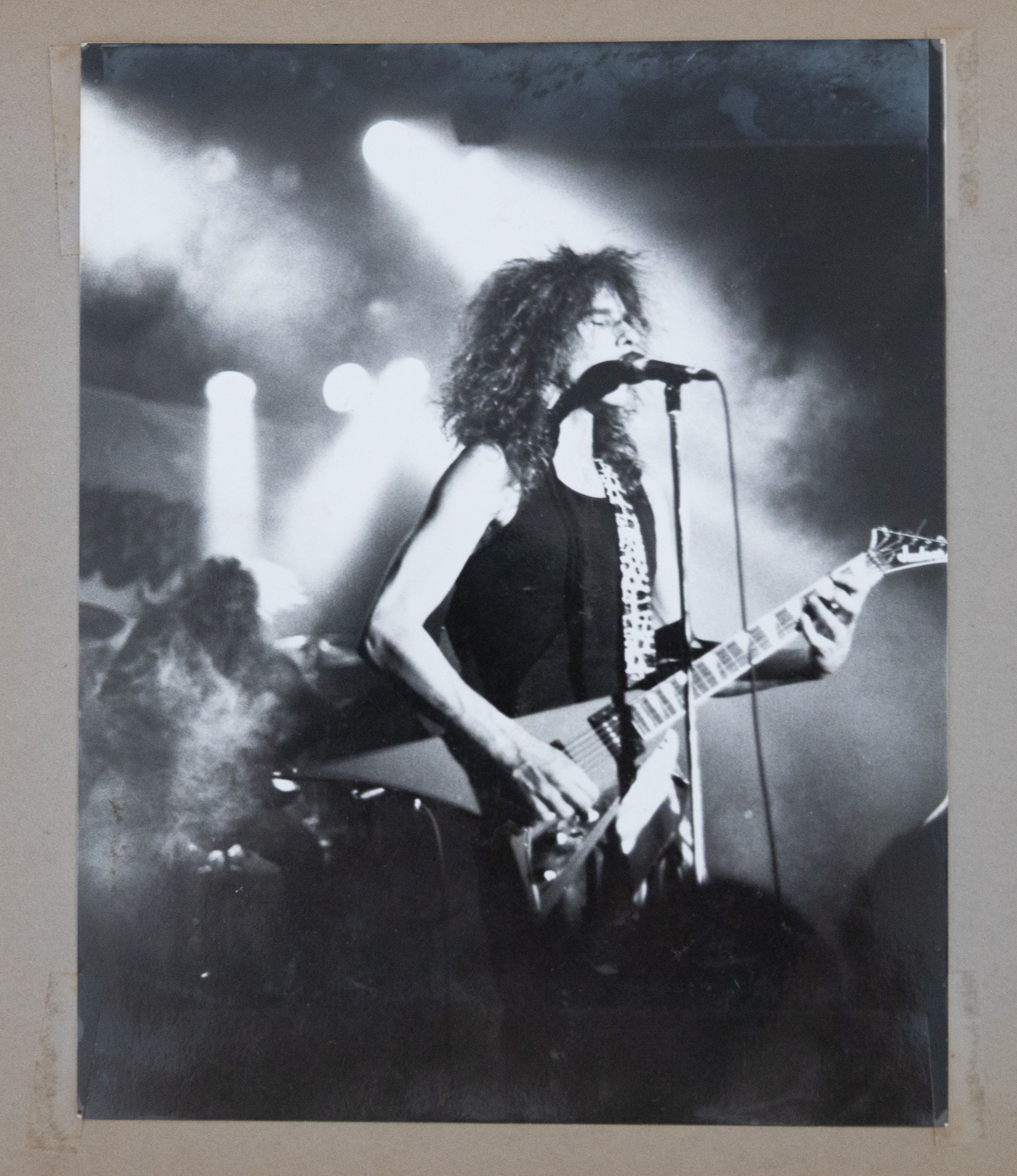 A black and white print mounted in a scrapbook showing Wally Passet singing into a microphone and playing a Flying V guitar under stage lights