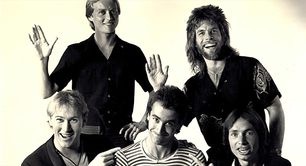 Black and white publicity portrait of five members of the band Bratz, smiling and waving at the camera