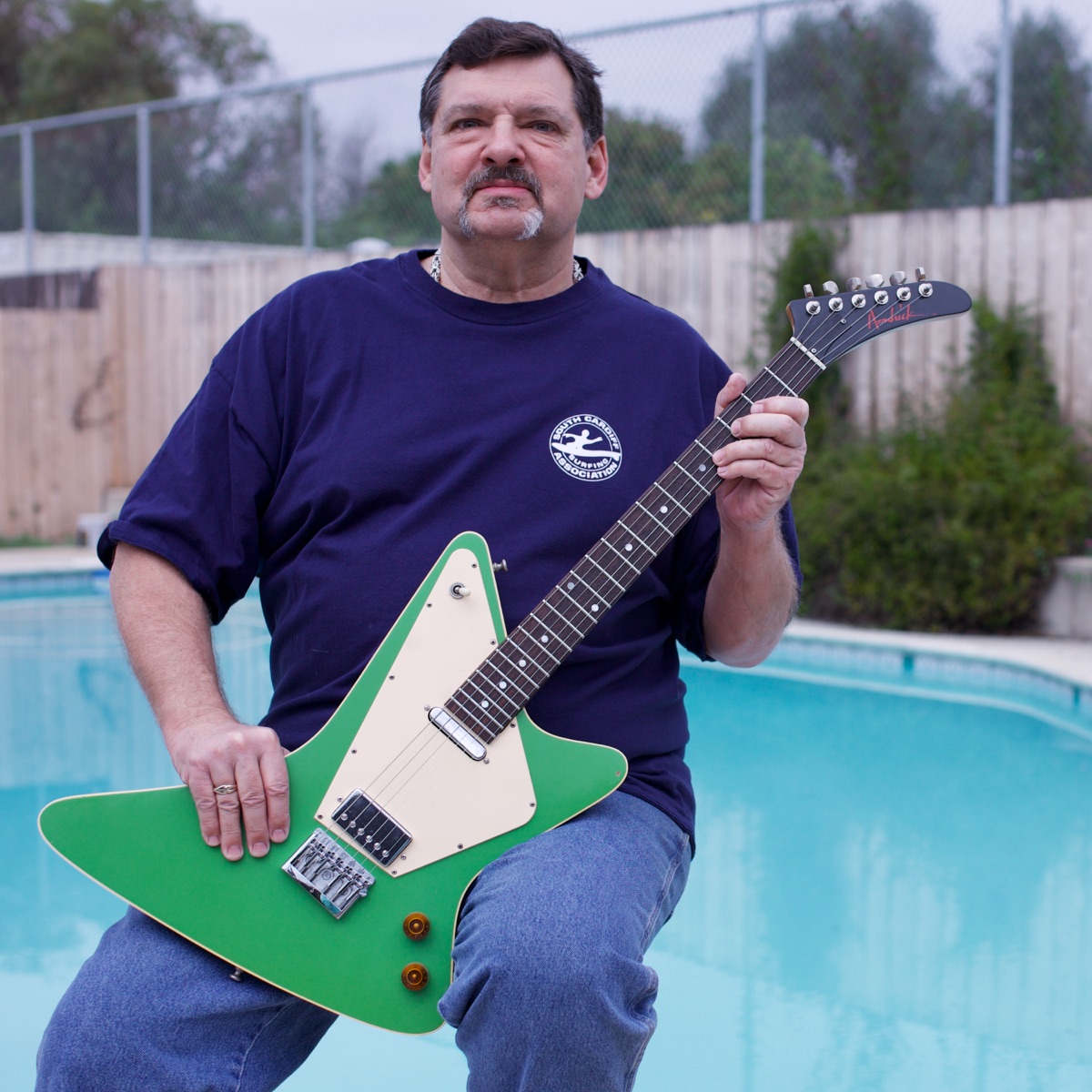 A man seated beside a swimming pool holding a bright green Hendrick Generator electric guitar with a cream pickguard