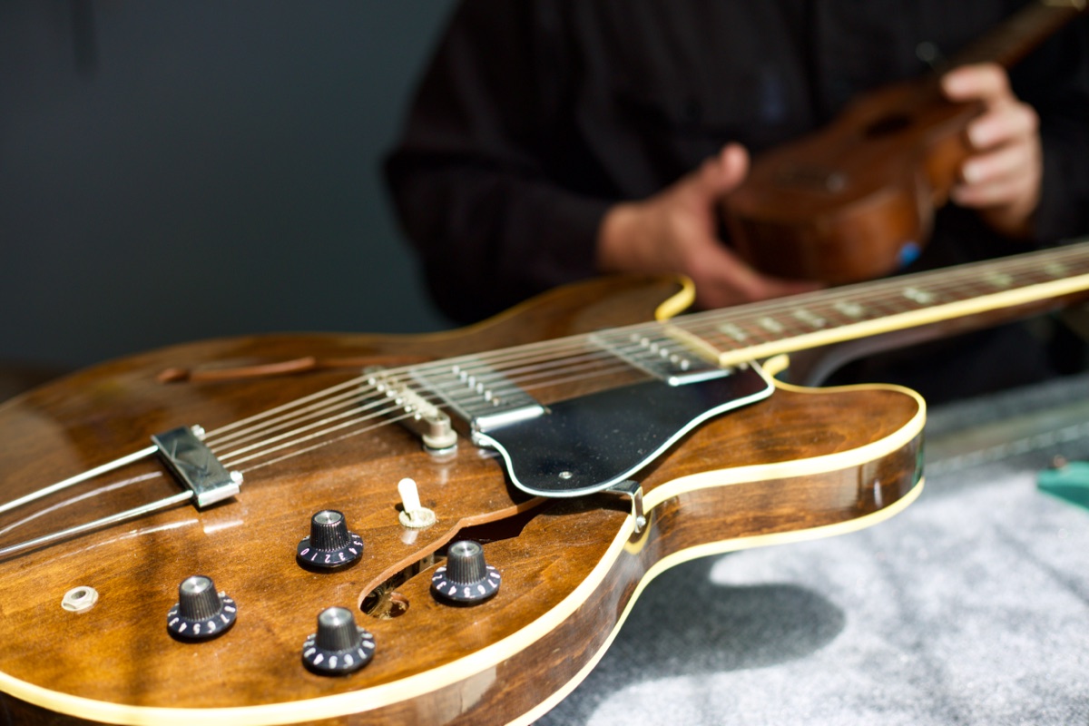 Close-up of a walnut-finished archtop electric guitar on the bench, knobs and pickup in focus, hands working on another guitar behind it