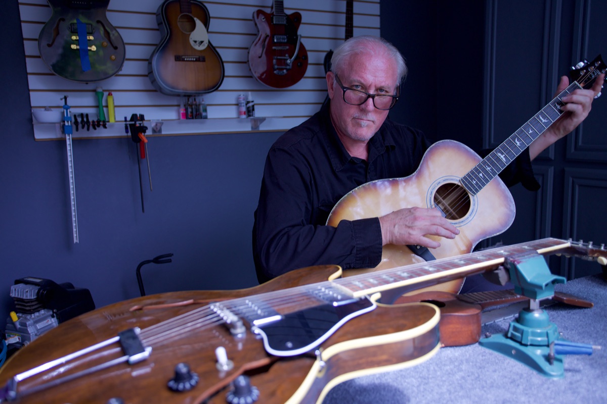 Lee Knight in his workshop holding a sunburst acoustic guitar, an archtop electric on the bench in front of him