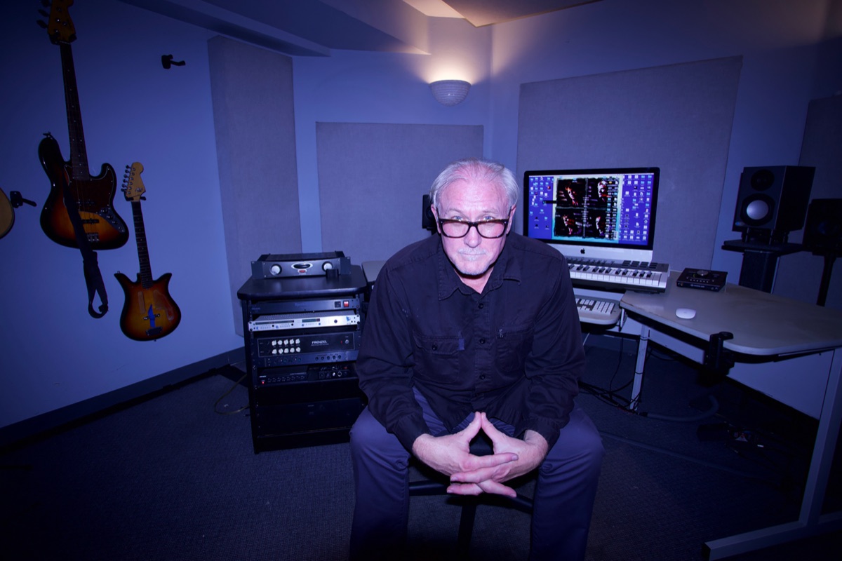 Lee Knight seated in his blue-lit recording studio, fingers steepled, rack gear and an iMac behind him, two basses on the wall