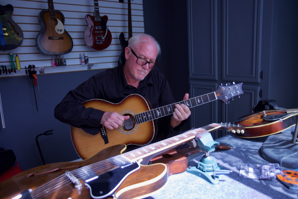 Lee Knight looking down as he tunes a sunburst acoustic guitar, guitars hanging on a slatwall behind him