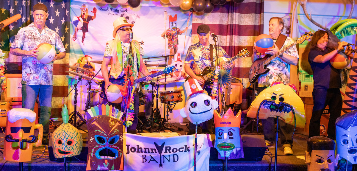 The Johnny Rock Band on stage at the Encinitas Elks Lodge surrounded by tiki heads, beach balls and hand-painted props, with a Johnny Rock Band banner across the front of the stage
