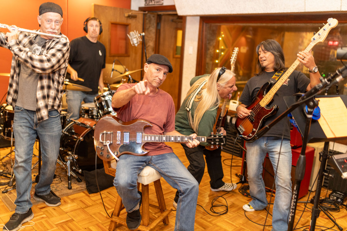 The Johnny Rock Band rehearsing indoors on a parquet floor, with flute, drums, lap steel and guitars