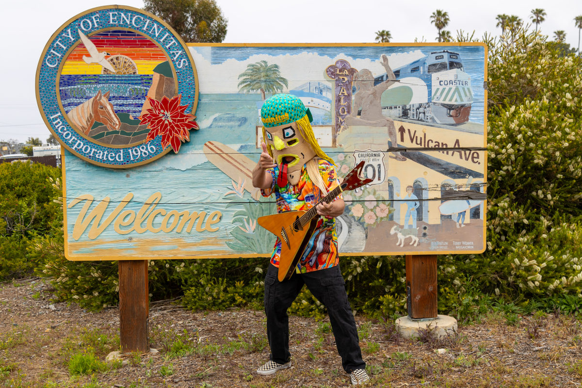 Tate Sanderson in mask and yellow wig playing an orange guitar in front of the City of Encinitas welcome sign on Vulcan Avenue