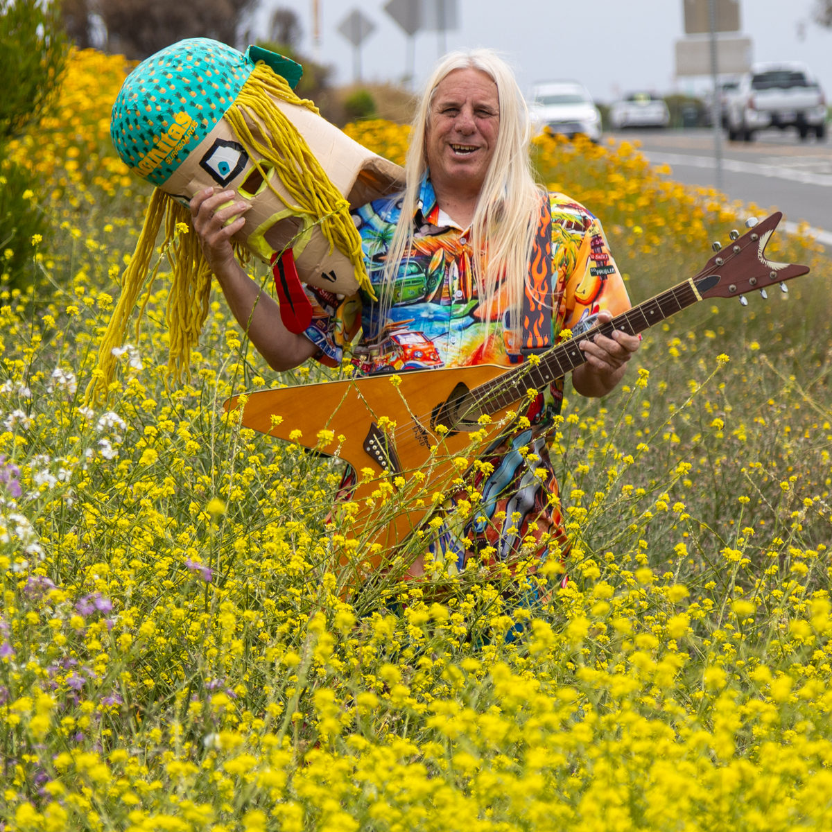 Tate Sanderson standing in a field of yellow mustard flowers beside Coast Highway, holding his mask and wig up beside his face and cradling an orange V-shaped guitar