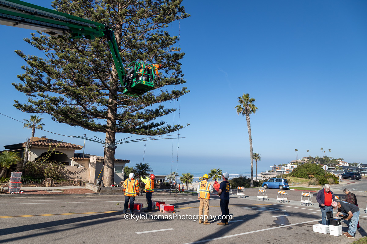 The street closed with cones and barricades, crew in orange high-visibility vests standing in the empty roadway, the boom basket raised into the branches above and a strand of lights angling down to the pavement