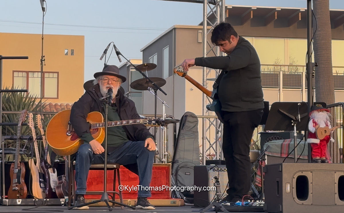 Jack Tempchin seated on stage in a dark hat with an acoustic guitar at the microphone, a second musician standing beside him tuning a guitar, amplifiers and a drum kit behind and a child in a Santa hat at the edge of the stage