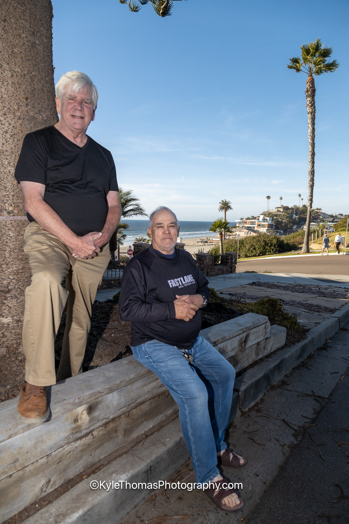 Jim Filanc standing and Luis Ortiz seated on the low sea wall above the beach, the ocean, palms and rooftops behind them on a clear day
