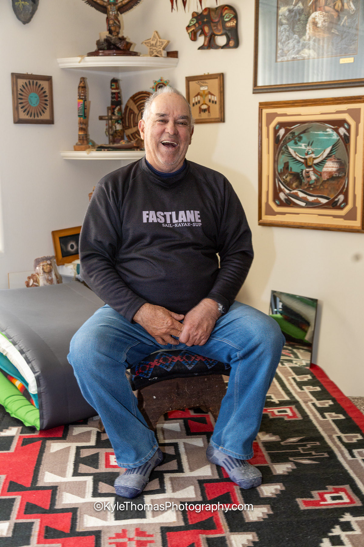 Luis Ortiz seated at home in a dark FASTLANE sweatshirt, laughing, the wall behind him covered with framed Native American art, carvings and keepsakes