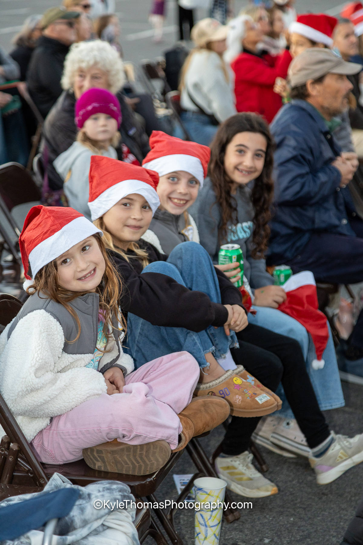Four girls in red Santa hats crowded together onto folding chairs, smiling at the camera, the packed crowd behind them