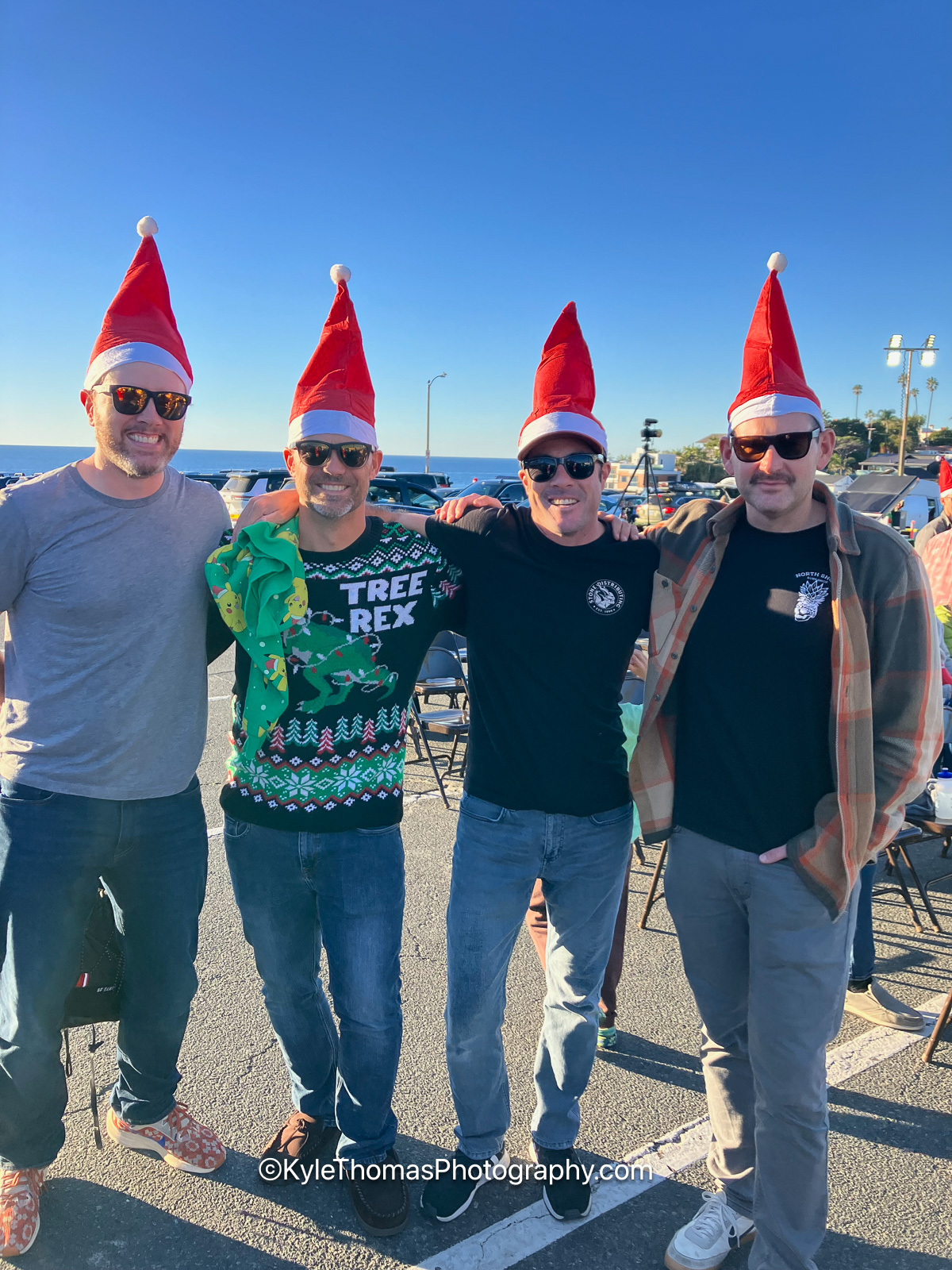 Four men in red Santa hats and sunglasses standing shoulder to shoulder and grinning, one in a green sweater reading Tree Rex, the ocean and parked cars behind them
