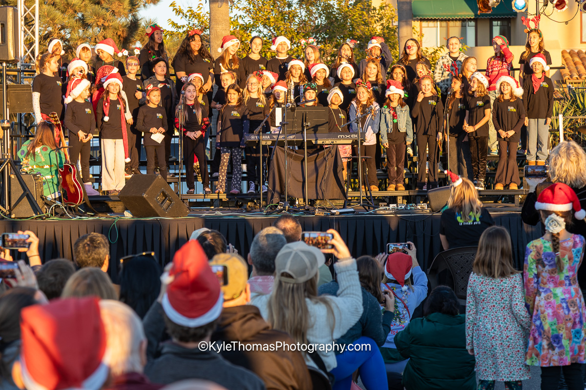 A school choir in black with Santa hats standing in rows on a lit stage behind keyboards and microphones, the audience in the foreground holding up phones to photograph them