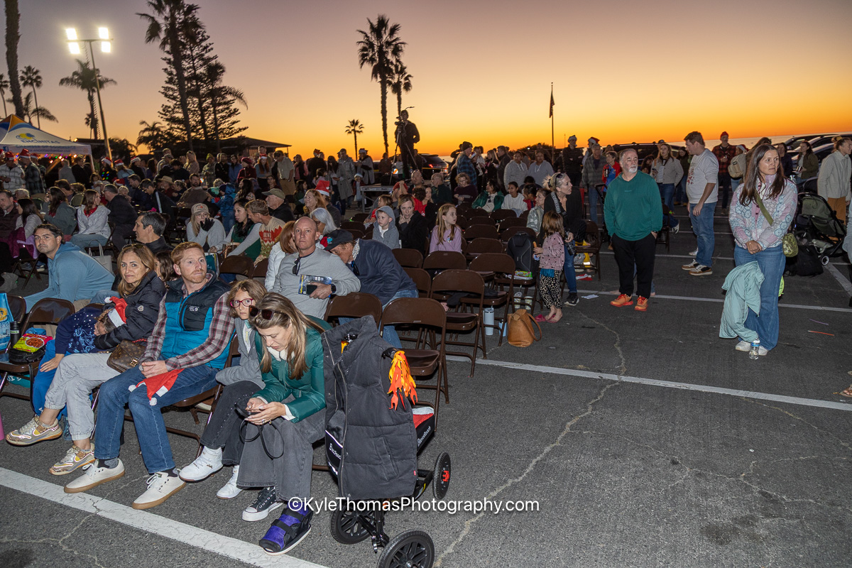 The seated crowd at dusk with an orange and gold sunset filling the sky over the ocean, palms and a flagpole in silhouette, people standing along the back of the lot