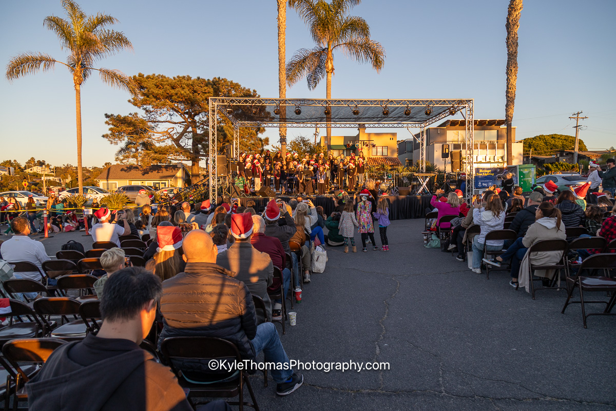 A wide view of a crowd seated on folding chairs in a parking lot facing a stage under lighting trusses, tall palms and rooftops behind, low golden sunlight across the pavement and Santa hats through the audience