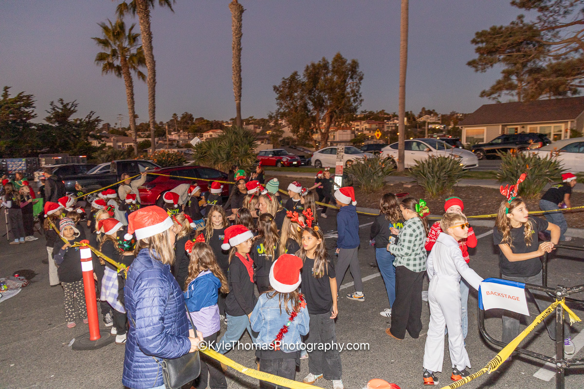A line of children in Santa hats, reindeer headbands and holiday sweaters waiting behind a yellow caution-tape rope and a Backstage sign, parents alongside them, palms and parked cars beyond